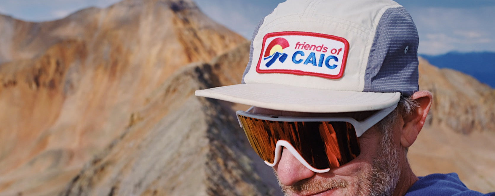 Person wearing a cap with 'Friends of CAIC' logo and sunglasses, set against a mountainous landscape.